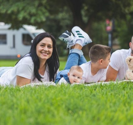 Happy family lying on grass with RV in the background, enjoying a sunny outdoor picnic.